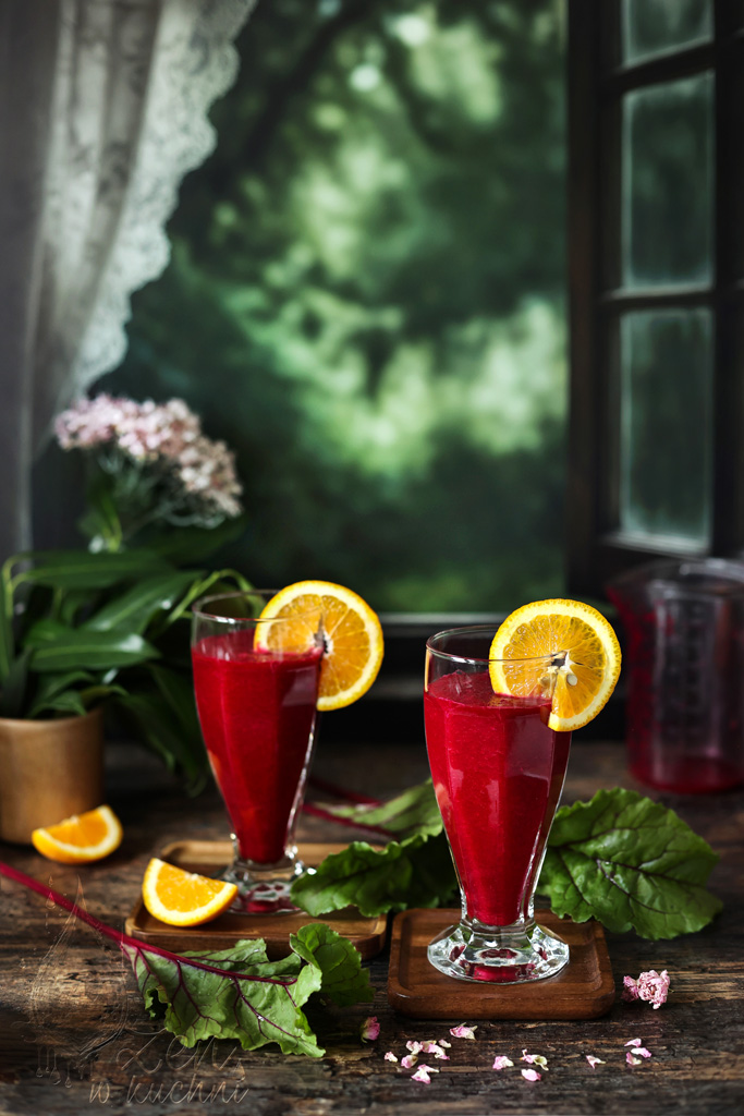 Dwie szklanki smoothie z buraków i pomarańczy na drewnianym blacie, obok plasterki pomarańczy i kawałki buraka